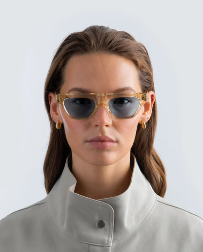 Woman wearing yellow sunglasses with blue lens and a beige coat on a light background