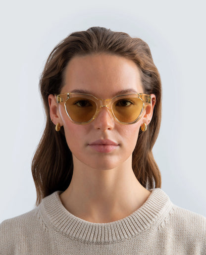 Woman wearing yellow sunglasses with yellow lens and a beige sweater against a light background