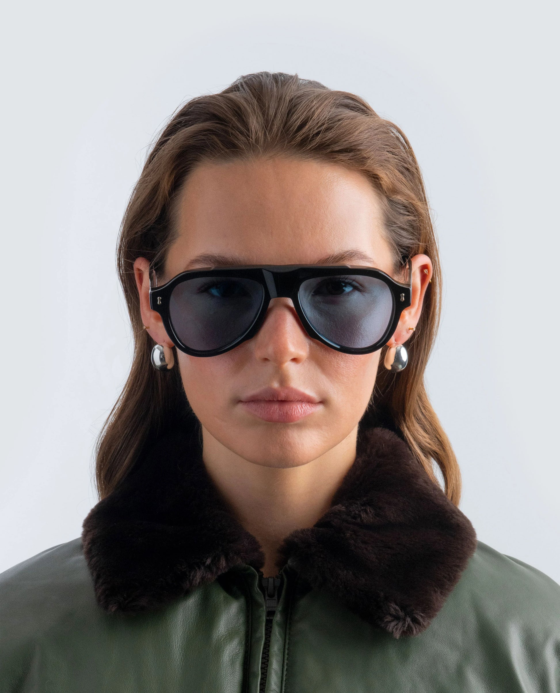 Woman wearing black sunglasses with blue lens and a green jacket against a light background