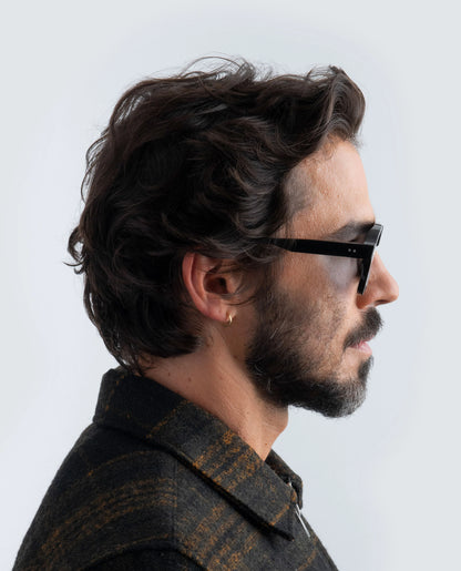 Side profile of a person with dark hair and beard wearing black sunglasses with blue lens against a light background