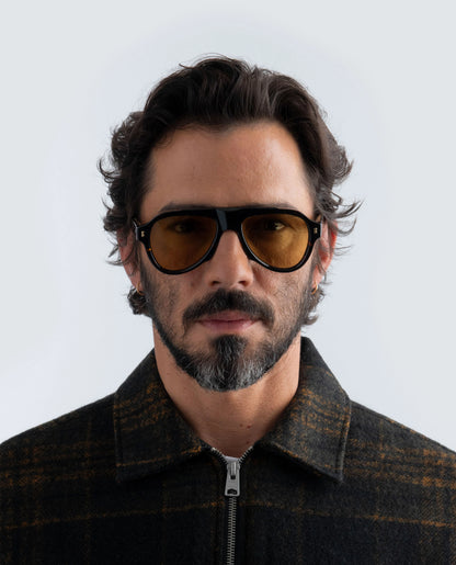 Man wearing Havana sunglasses with amber lens and a dark jacket against a light background