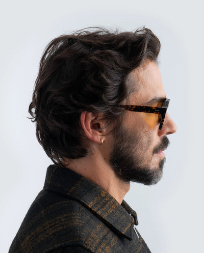 Man wearing Havana sunglasses with amber lens and a dark jacket against a light background