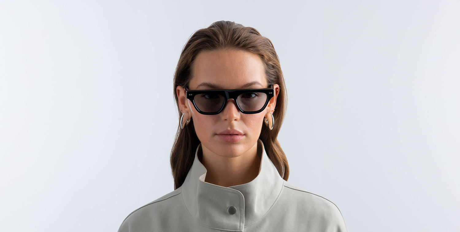 Woman wearing black sunglasses with gray lenses and a light gray jacket on a white background