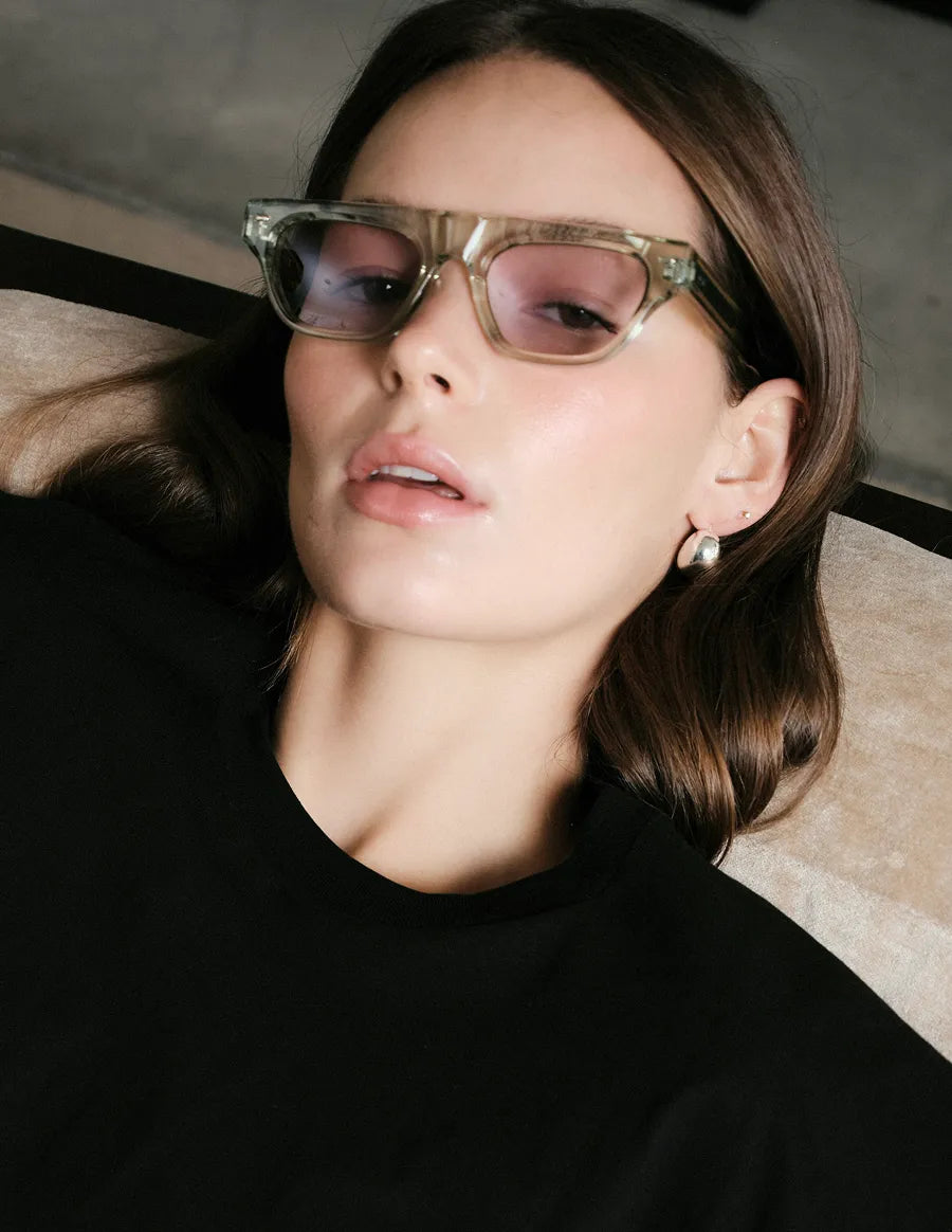 Woman wearing seafoam green sunglasses with lilac lenses and a black top in an indoor setting