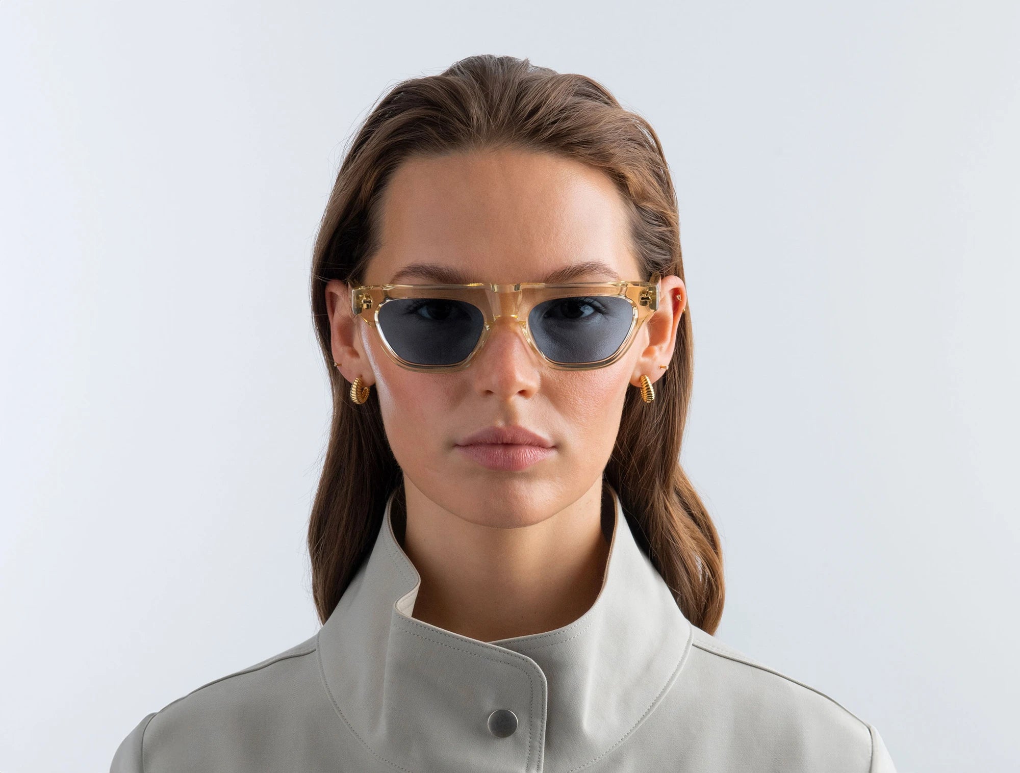 Woman wearing yellow sunglasses with blue lenses and a light gray jacket against a white background