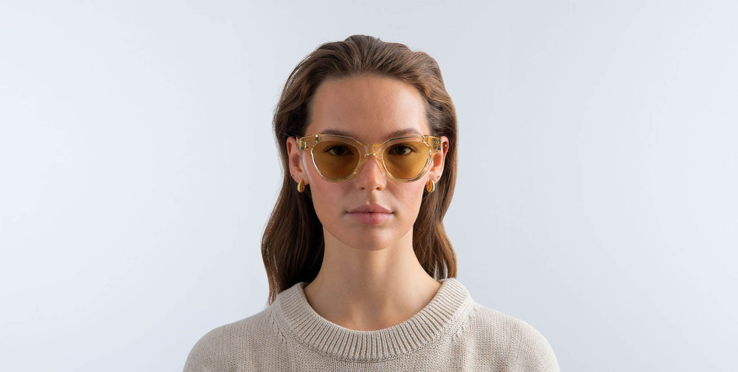 Woman wearing champagne yellow sunglasses with yellow lenses against a light gray background