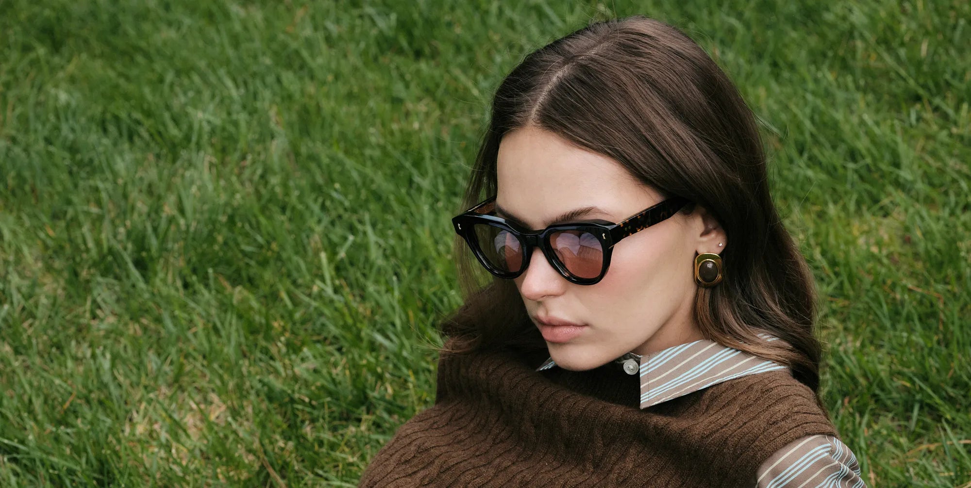 Woman wearing tortoiseshell sunglasses  with brown lenses and a brown sweater in a grassy outdoor setting