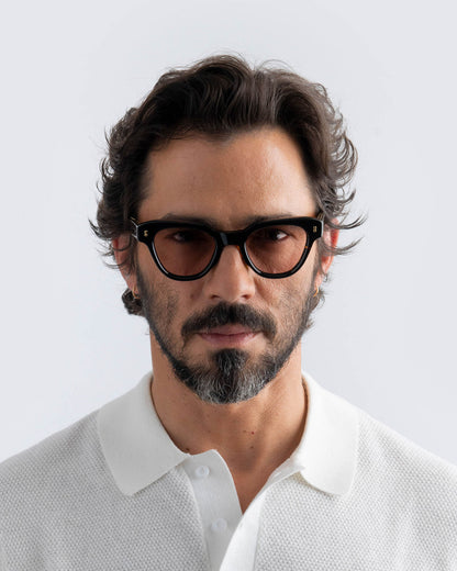 Man wearing tortoiseshell cat-eye sunglasses with brown lenses and a white shirt against a light gray background