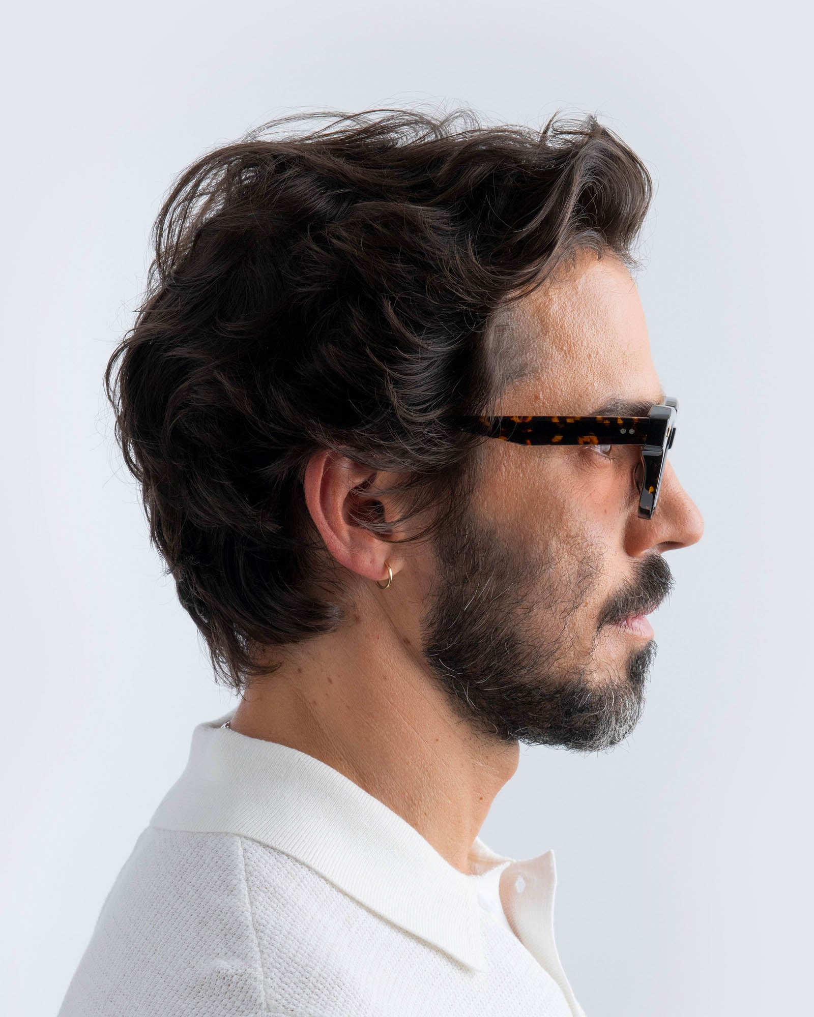 Man wearing tortoiseshell cat-eye sunglasses with brown lenses and a white shirt against a light gray background
