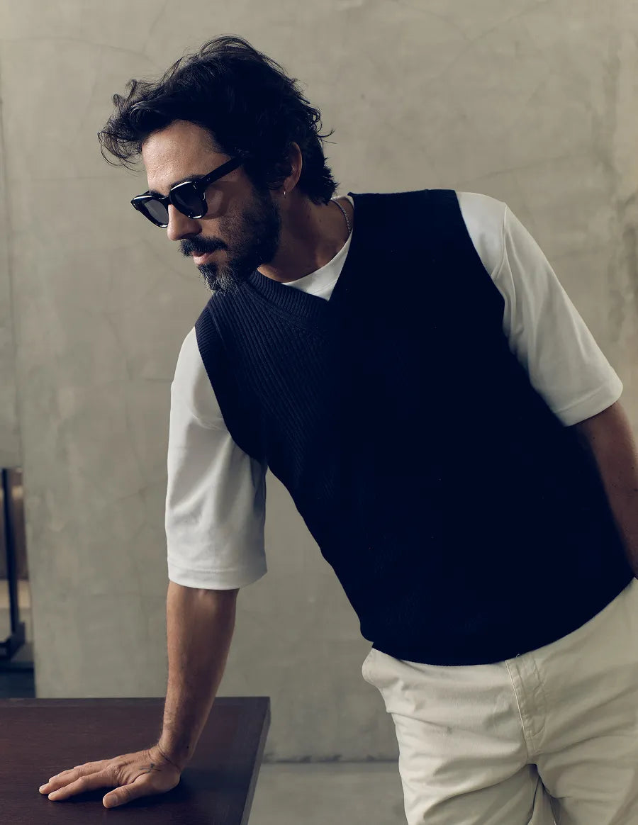 Man wearing black sunglasses with black lenses and a dark vest over a white shirt against a neutral background