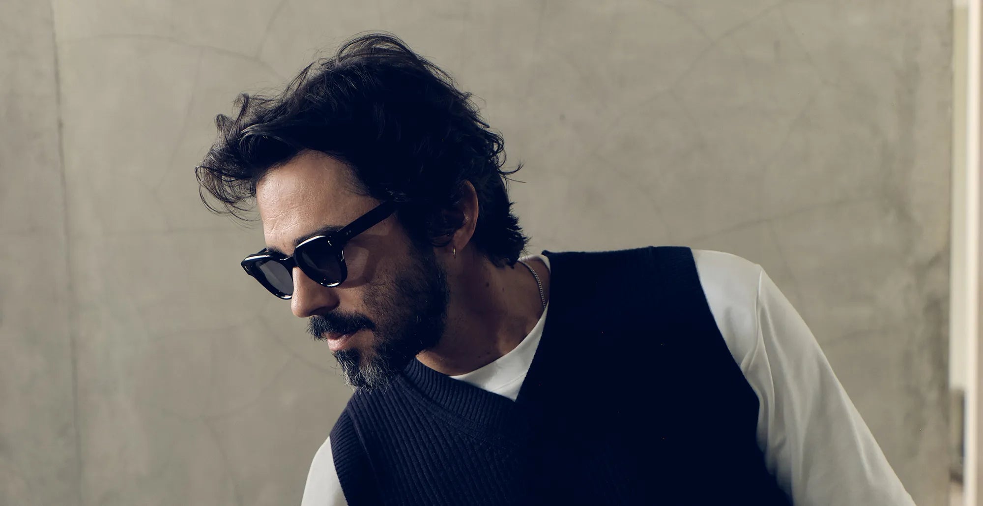 Man wearing black sunglasses with black lenses and a dark vest over a white shirt against a neutral background