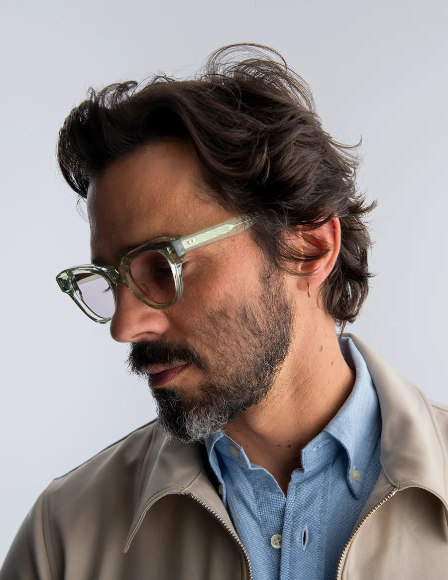 Man wearing seafoam green sunglasses and a beige jacket with a light blue shirt against a gray background
