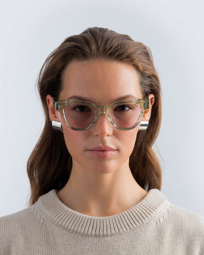 Woman wearing seafoam green sunglasses with lilac lenses against a light grey background