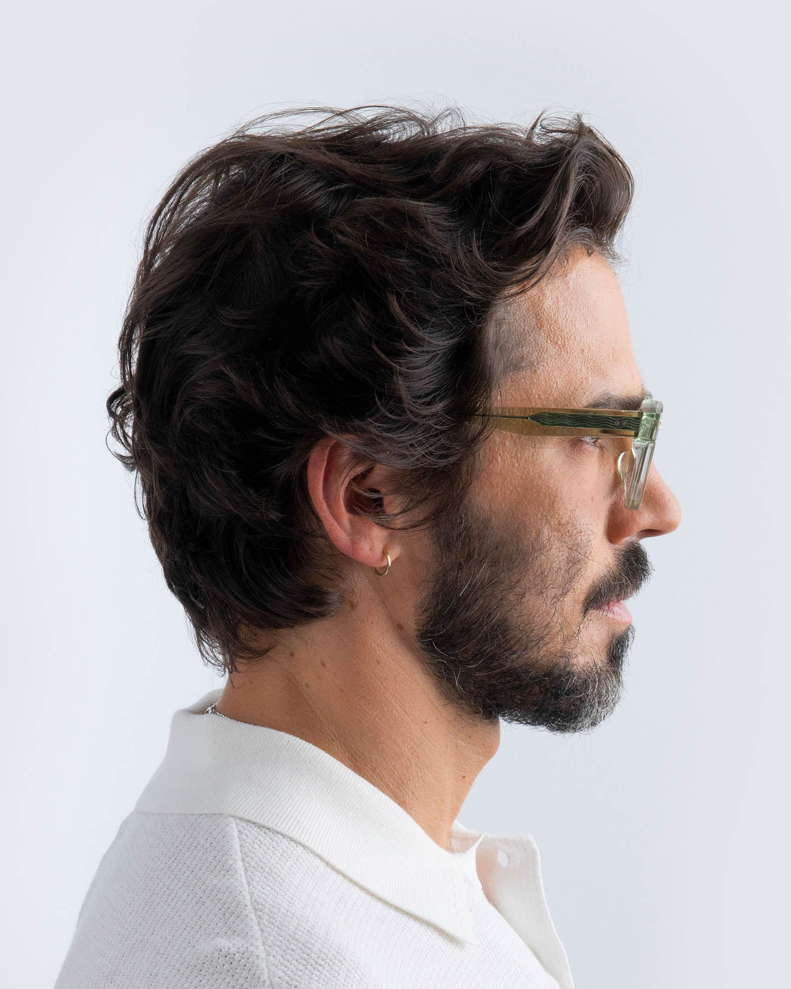 Man with dark hair and beard wearing seafoam green sunglasses with lilac lenses and a white shirt on a light background