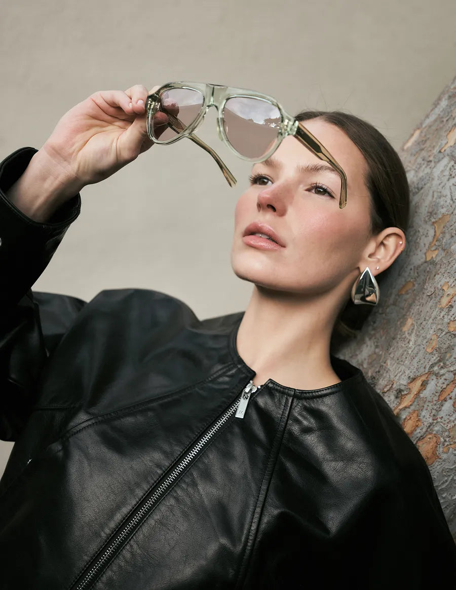 Woman holding a pair of seafoam green sunglasses with lilac lenses up to her face against a neutral background