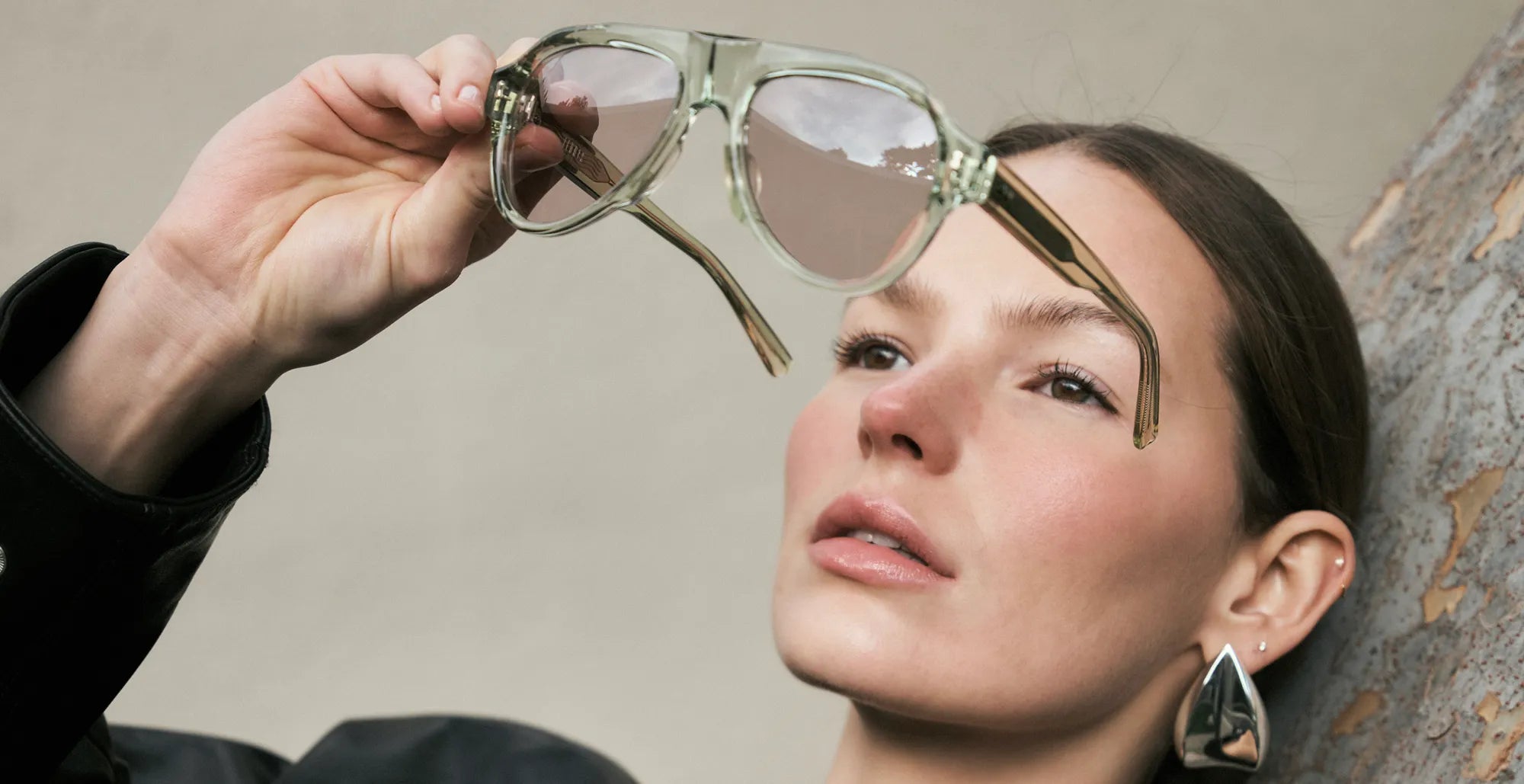 Woman holding a pair of seafoam green sunglasses with lilac lenses up to her face against a neutral background