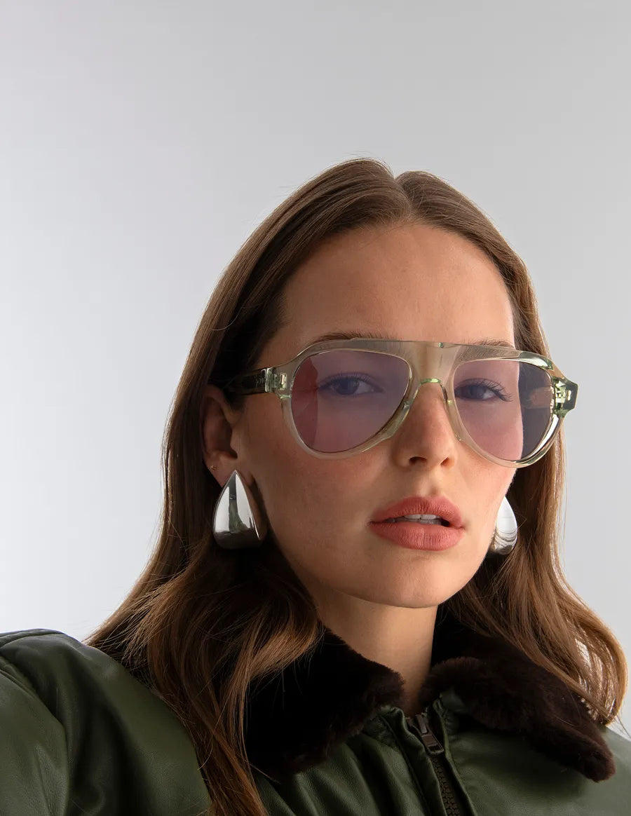 Woman wearing aviator style sunglasses with seafoam green frames and lilac lenses with a plain background