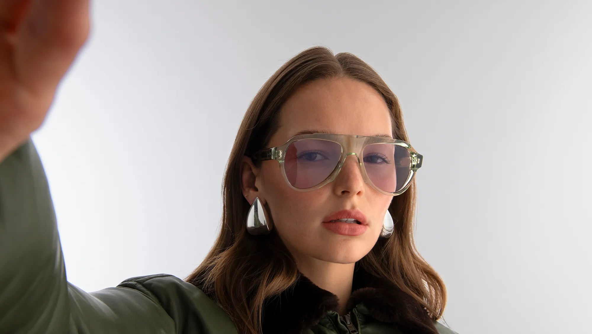 Woman wearing aviator style sunglasses with seafoam green frames and lilac lenses with a plain background