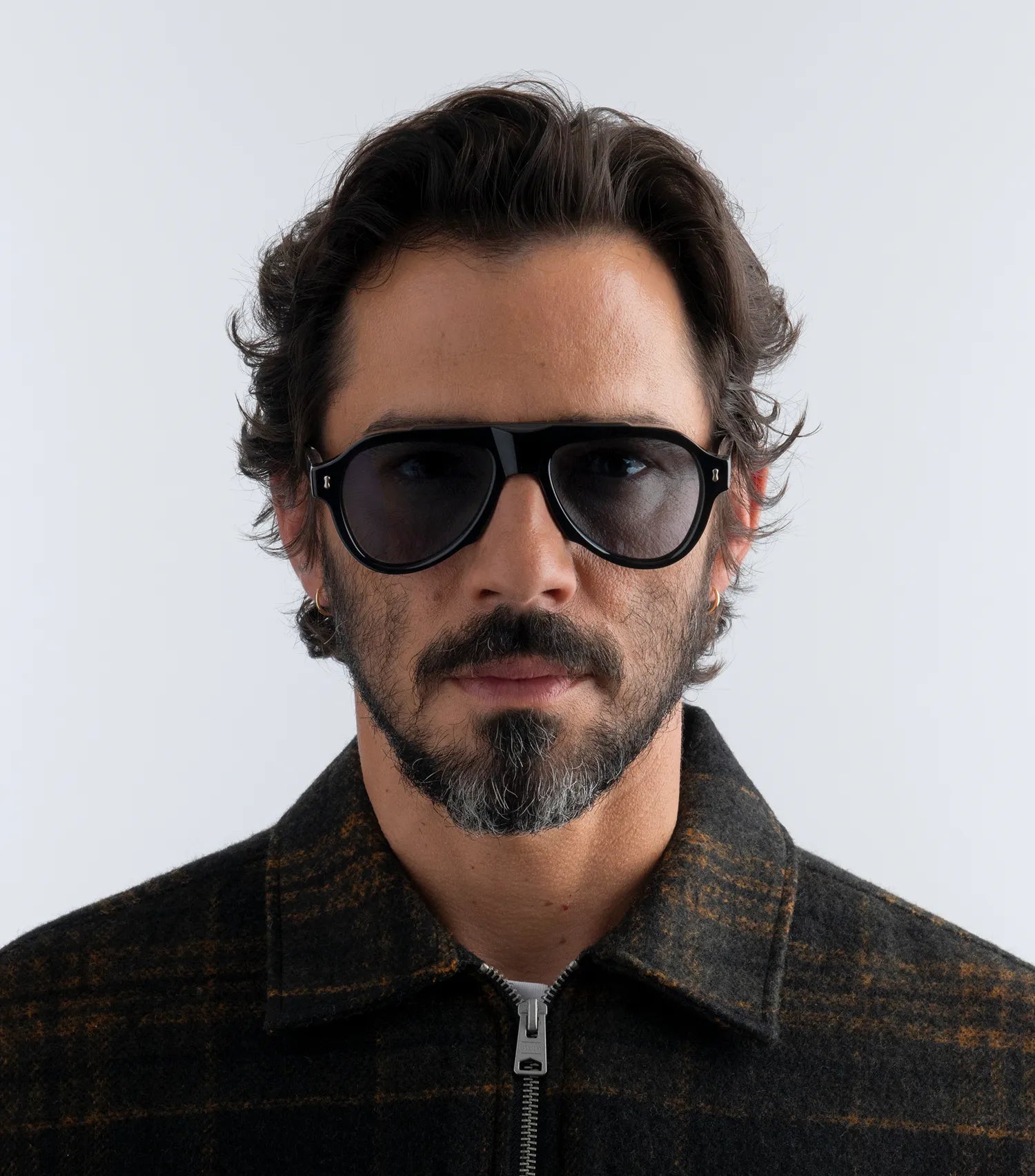 Man wearing black aviator style sunglasses with black lenses and a dark jacket against a plain background