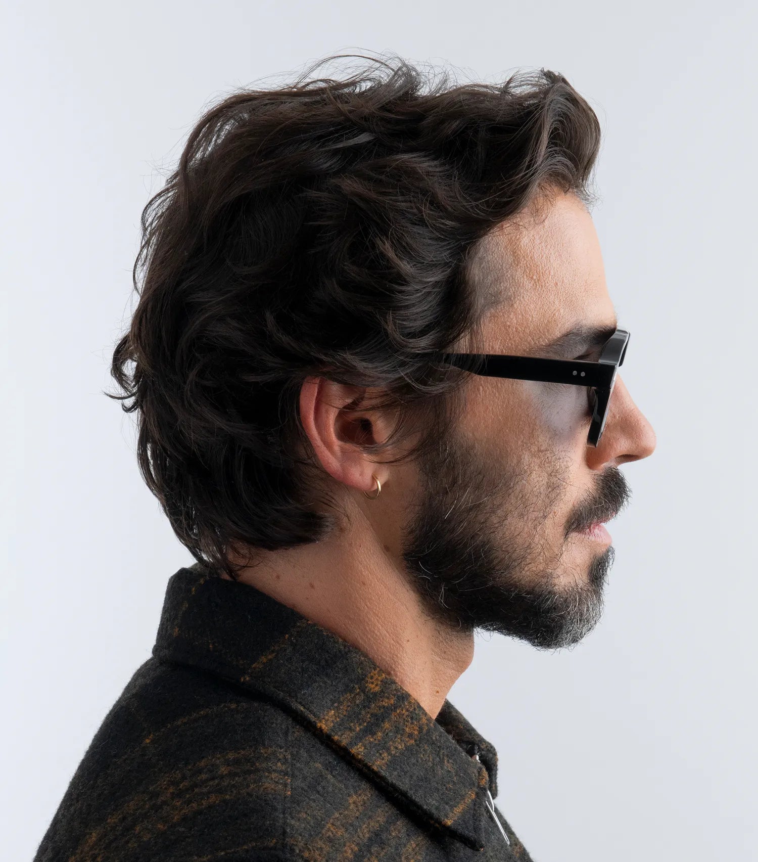 Man wearing black aviator style sunglasses with black lenses and a dark jacket against a plain background