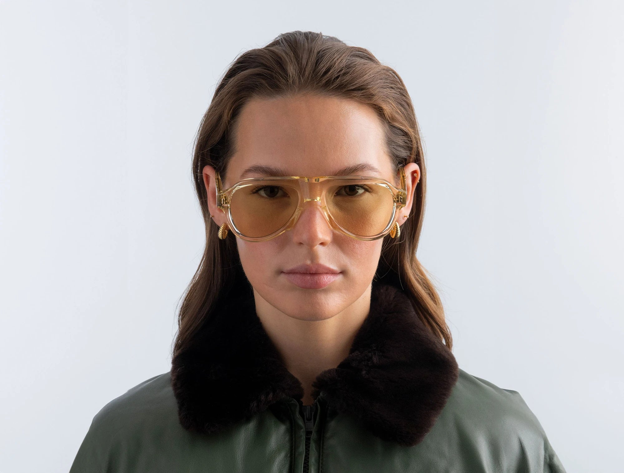 Woman wearing yellow aviator-style sunglasses with yellow lenses and a green coat with a fur collar against a white background
