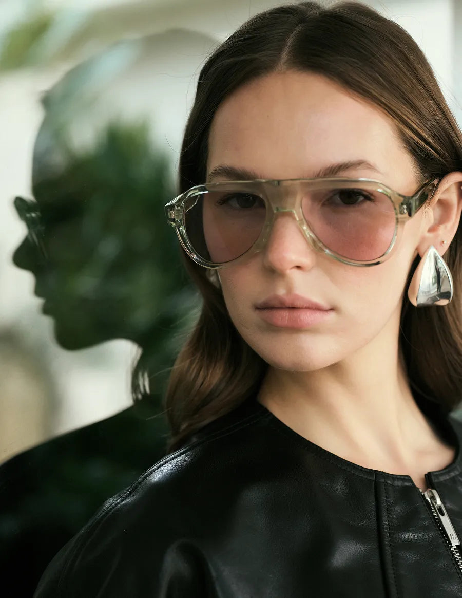 Woman wearing clear seafoam green sunglasses and a black leather jacket with a blurred green plant background